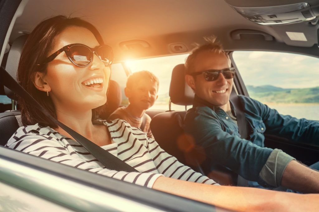 Smiling family in a car wearing seatbelts, enjoying a sunny drive, highlighting safe and comfortable travel.
